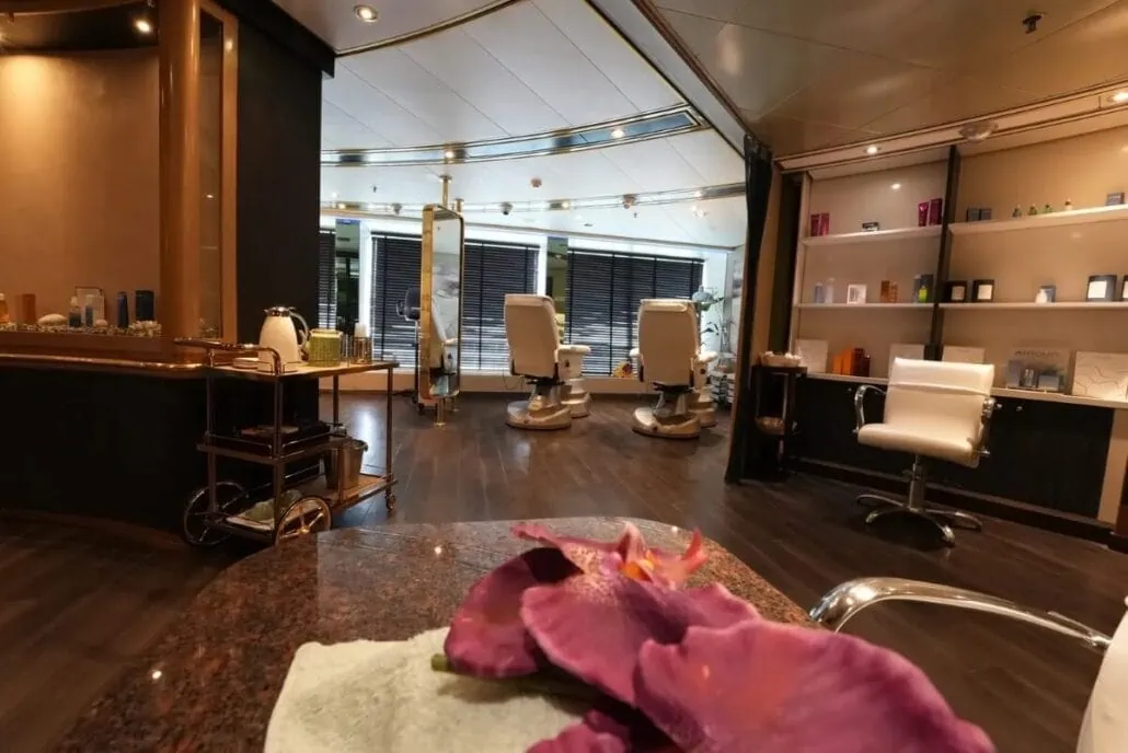 A modern salon with two styling chairs facing large windows offers a serene view reminiscent of the Arabian Gulf itinerary. Shelves line the right wall, displaying various products. A table in the foreground holds purple flowers and a towel, all elegantly illuminated by overhead lighting.