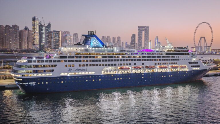 A large cruise ship, part of the renowned Celestyal Cruises, docked at a port with its logo proudly displayed. In the background, a city skyline with tall buildings and a ferris wheel stretches under a twilight sky, hinting at an exciting itinerary ahead.