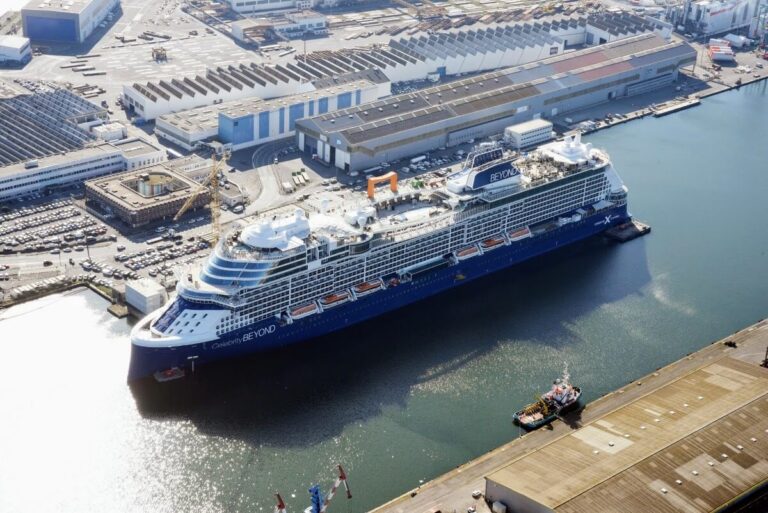 A majestic cruise ship, reminiscent of the Celebrity Cruises Edge Series, docked at a bustling industrial port. The blue and white vessel towers above warehouses, parked vehicles, and a tugboat near the water's edge.