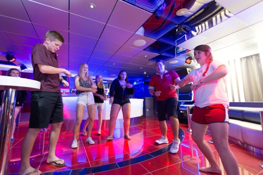 A group of six people are dancing energetically in a brightly lit room with red flooring and white walls, evoking a lively carnival vibe. They are casually dressed, enjoying the moment. In the background, a bar counter and stools suggest stricter rules for guests under 21.