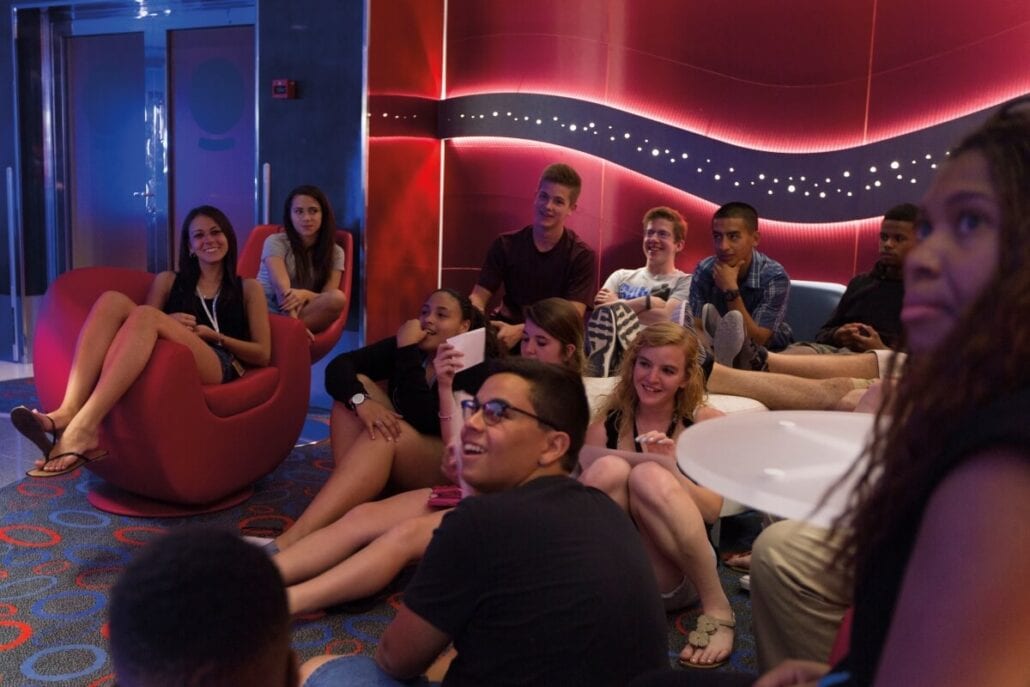 A group of guests under 21 are seated on the floor and in chairs in a modern room with red and blue lighting. They appear to be watching something, with some smiling and others focused. The room has circular patterns on the carpet, reminiscent of a vibrant Carnival atmosphere.