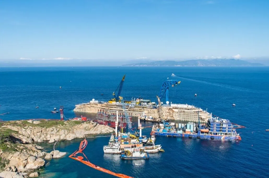 Aerial view of a large shipwreck reminiscent of the Costa Concordia disaster near the coastline, with cranes and vessels working on the site. The ship is partially submerged in the clear blue sea, with orange containment booms surrounding the area. Rocky coastline visible in the foreground.