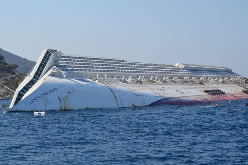 The image depicts the Costa Concordia, a large white cruise ship disaster partially submerged and tilted on its side near a rocky shore. The ship's name is visible on the hull. Despite the calamity, the sky remains clear and the water appears calm.