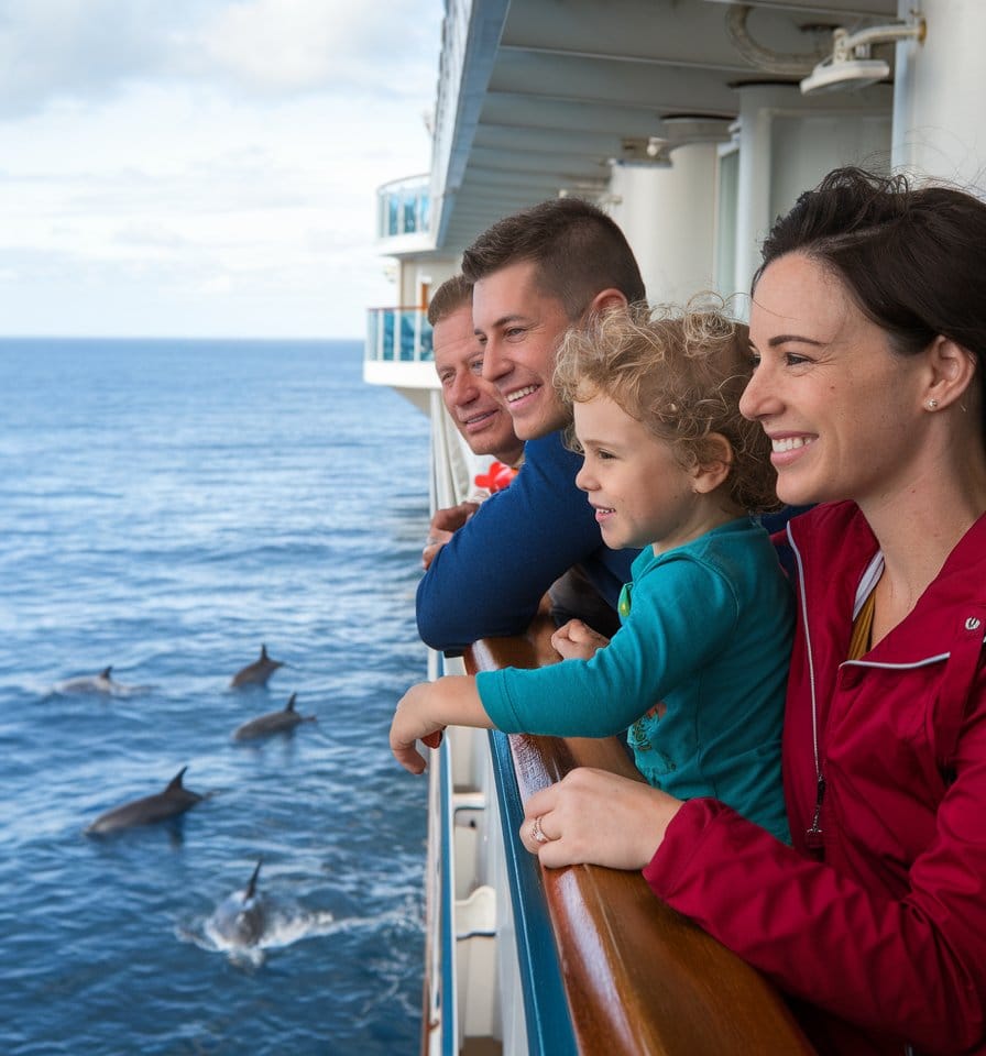 A family cruise offers endless fun photo ideas as a group, including a young child, stands on the ship's deck. They watch in awe as dolphins play in the ocean below, all while leaning over the wooden railing against a clear sky backdrop—perfect for capturing memorable cruise photography.