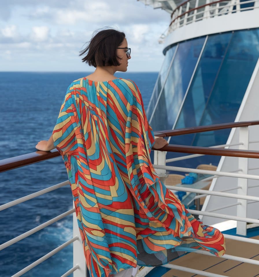 A person with short hair stands on a cruise ship deck, gazing at the ocean in February. Sporting one of those must-have tropical cruise outfits, they wear a colorful, flowing dress adorned with vibrant orange, blue, and yellow patterns. The sky above is partly cloudy.