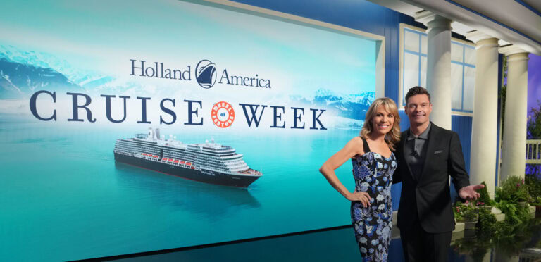 Two people stand in front of a backdrop displaying "Holland America Cruise Week" with an image of a cruise ship and snowy mountains. Smiling and posing for a photo, they celebrate the exciting Wheel of Fortune partnership that offers lucky winners the chance to enjoy free cruises.
