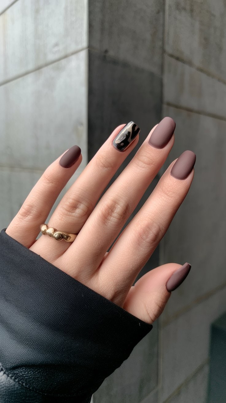 A chic hand with matte brown nails is shown, with one nail flaunting a striking leopard print design. The person wears a black sleeve and a gold ring on their ring finger, capturing the essence of sophisticated short nail designs. The background is a minimalist concrete wall.