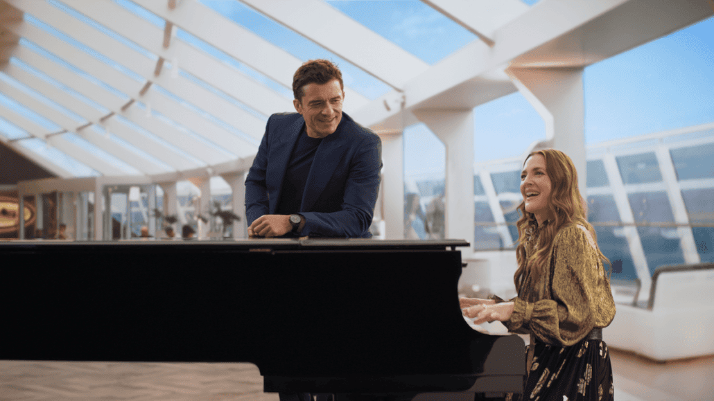 A man in a blue suit leans on a black grand piano while a woman in a patterned blouse, reminiscent of Orlando Bloom’s casual elegance, plays it. They are in a modern, well-lit room with large windows and white furniture. Both appear to be smiling and enjoying the moment.