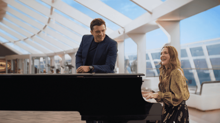 A man in a blue suit leans on a black grand piano while a woman in a patterned blouse, reminiscent of Orlando Bloom’s casual elegance, plays it. They are in a modern, well-lit room with large windows and white furniture. Both appear to be smiling and enjoying the moment.