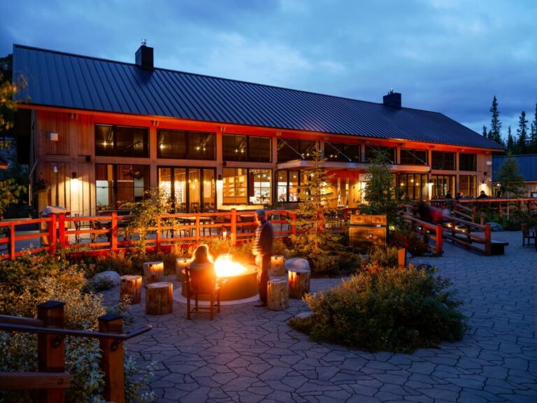 A rustic lodge with large windows is warmly lit at dusk, offering quintessential Alaskan experiences. In the foreground, people gather around a glowing fire pit surrounded by wooden stumps and greenery on a stone patio. Towering trees are visible in the background, enhancing the serene atmosphere.
