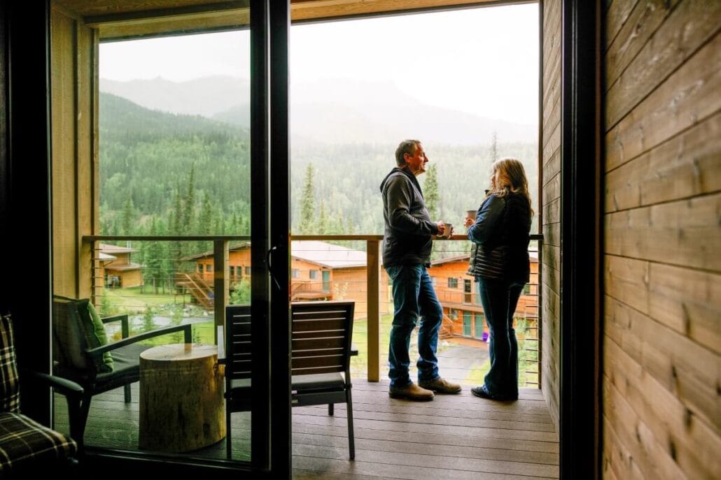 Two people stand on a wooden balcony, savoring their Alaskan experience with mugs in hand. They gaze at the lush green trees and mountains framed by an open glass door. Chairs and a small table invite relaxation, much like the tranquil cruises offered by Holland America.
