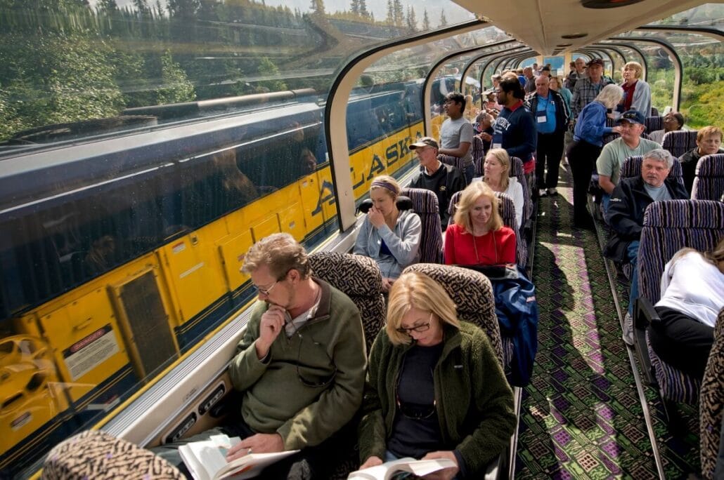 Passengers are seated inside a train with large windows, some reading and others enjoying the Alaskan experience. A yellow and blue train with "Alaska" written on it, reminiscent of Holland America's $70M expansion, is visible through the windows. Trees and a landscape can be seen in the background.