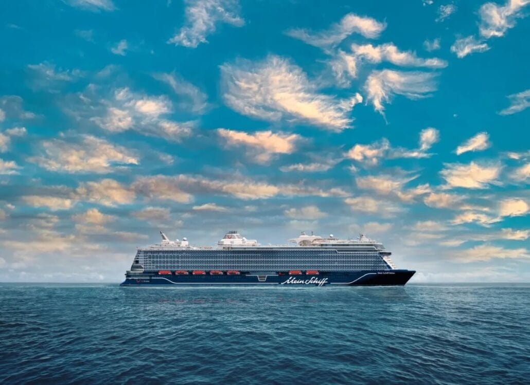 A large, modern cruise ship from the new 2025 fleet sails on calm waters under a sky with scattered clouds. Painted dark blue with a logo on the side, the ship harmoniously moves along the horizon, separating sea from sky and creating a serene scene.