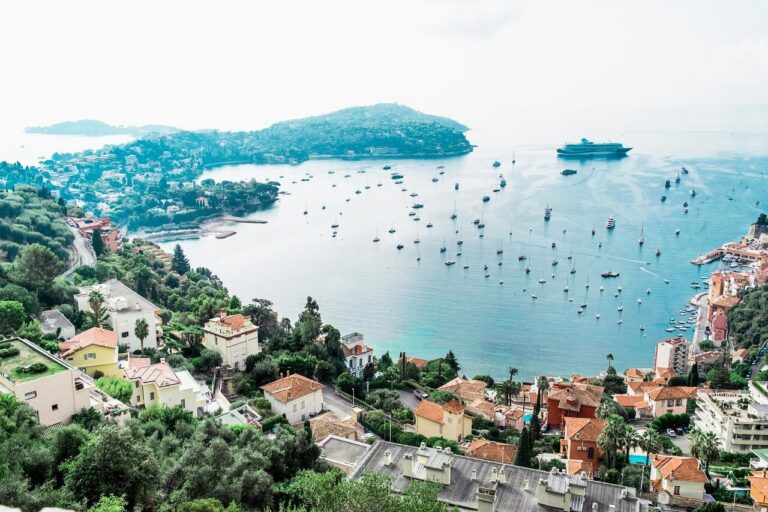 A scenic coastal view of Nice, France, reveals a town with red-roofed buildings and lush greenery. The bay, unaffected by the cruise ship ban, is dotted with numerous small boats and a large cruise ship. Hills surround the water under an overcast sky filled with soft light.