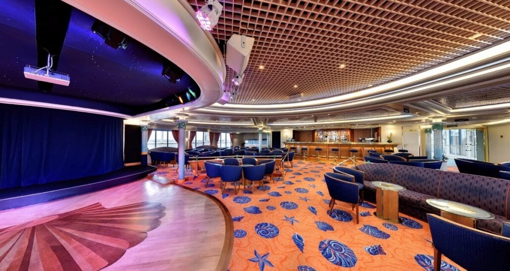 A modern, spacious lounge at Villa Vie Residences welcomes you with an orange carpet adorned with blue floral patterns. Blue chairs and tables are arranged near a stage with a dark blue curtain, while the ceiling's grid design features ambient lighting. A bar stands visible in the background.