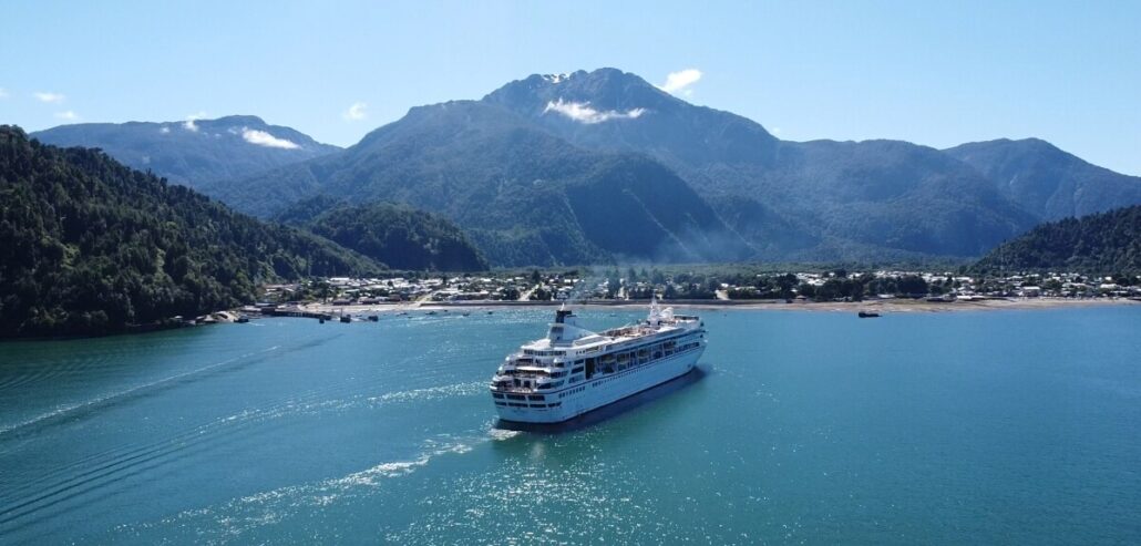 A cruise ship majestically sails on a blue body of water with Villa Vie Residences nestled in the coastal town against a backdrop of lush, green mountains. Under the clear blue sky, this idyllic scene invites thoughts of seasonal ownership and serene escapes.