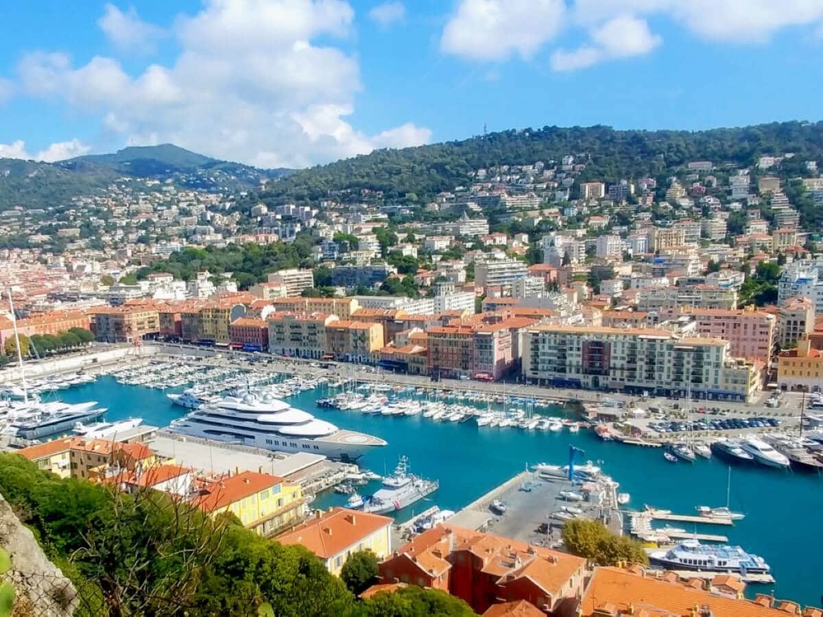 A coastal city reminiscent of Nice, France, boasts a marina with numerous boats and a large yacht, surrounded by red-roofed buildings. Hills lush with greenery rise in the background under a partly cloudy sky, free from large cruise ships due to a recent ban.