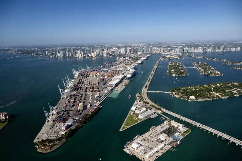 Aerial view of a bustling port area with cranes and cargo containers on an island, connected by bridges to the mainland city skyline. Amidst turquoise waters and small residential islands, law enforcers work diligently to prevent smuggling narcotics. Clear blue sky above.