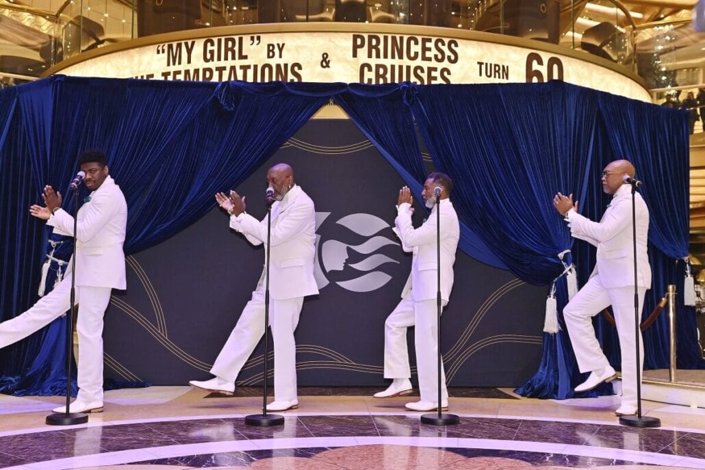 Four men in white suits, embodying the classic charm of a Motown singing group, perform on a stage with microphones. In front of a dark blue curtain inside a venue, the backdrop celebrates "My Girl" and "Princess Cruises," as they strike coordinated poses with one leg raised.
