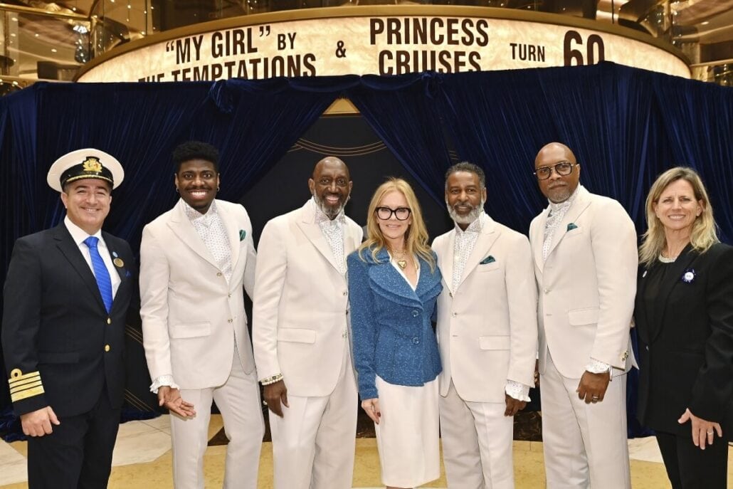 A group of seven people, including four men in light-colored suits, two women in business attire, and a man in a captain's uniform, stand smiling in front of a navy blue curtain. The background features a sign celebrating Princess Cruises' 60th anniversary with echoes of Motown vibes.