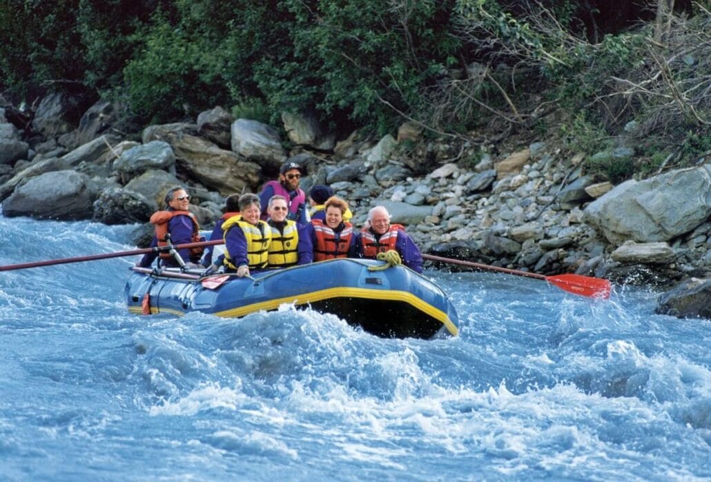 A group of adventurers wearing life jackets navigate a raft through whitewater rapids, experiencing the thrill akin to Alaskan Experiences. The river is flanked by rocky banks and dense greenery, with an oar clutched in the grip of one individual.