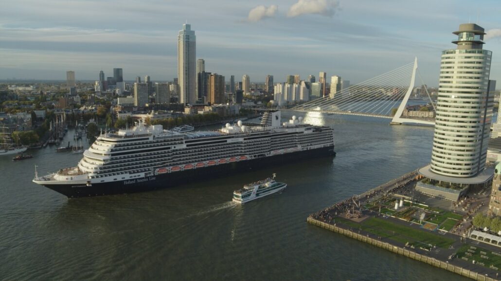 A large cruise ship, reminiscent of the luxurious Holland America line, navigates a river with a smaller boat nearby. The cityscape is adorned with modern skyscrapers and a striking white cable-stayed bridge, all under a partly cloudy sky.