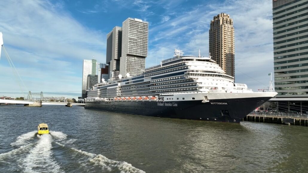 A large cruise ship, named Rotterdam, from Holland America is docked beside a city waterfront with tall buildings. A small boat glides through the water in the foreground under a partly cloudy sky, reminiscent of winning free cruises on Wheel of Fortune.