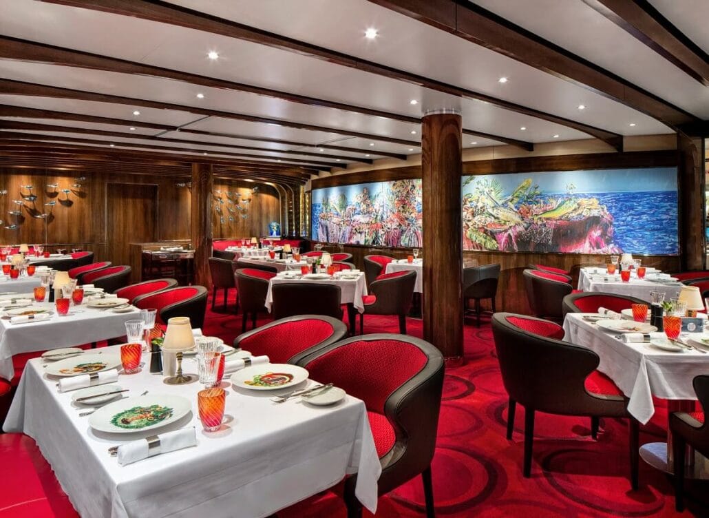 Elegant Mediterranean bistro interior with white tablecloths, red cushioned chairs, and wood accents. Tables are set with plates, glasses, and cutlery. A large, colorful mural is visible on the wall of Rudi’s Sel de Mer on Holland America, and the ceiling features recessed lighting.
