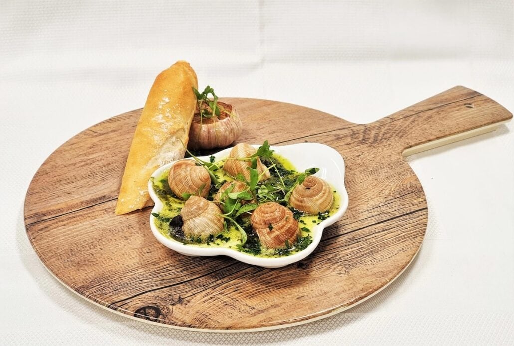 A wooden serving board presents a dish of escargot with garlic butter and herbs at Holland America's Rudi’s Sel de Mer. A baguette slice accompanies the arrangement, set against a white backdrop that draws attention to this Mediterranean Bistro delight.