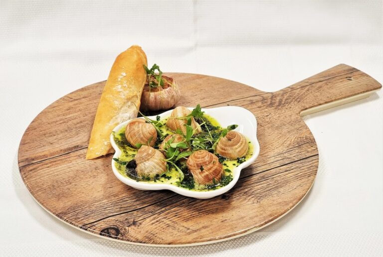 A wooden serving board presents a dish of escargot with garlic butter and herbs at Holland America's Rudi’s Sel de Mer. A baguette slice accompanies the arrangement, set against a white backdrop that draws attention to this Mediterranean Bistro delight.