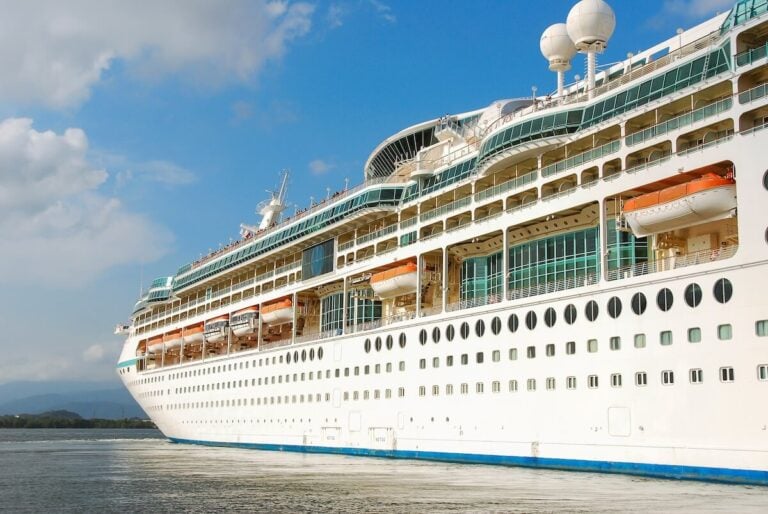 A large white Royal Caribbean vessel is docked at a port, its multiple decks lined with orange lifeboats. The sky is clear with a few clouds, and the sea is calm.