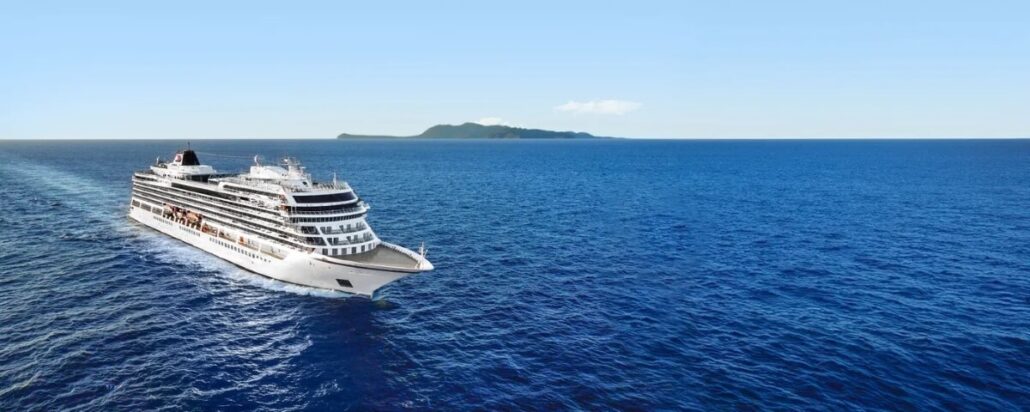 A large white cruise ship, one of the new ships in the cruise industry, sails on a vast, calm blue ocean. An island is visible on the horizon under a clear 2025 sky. The ship is moving left to right in the frame.