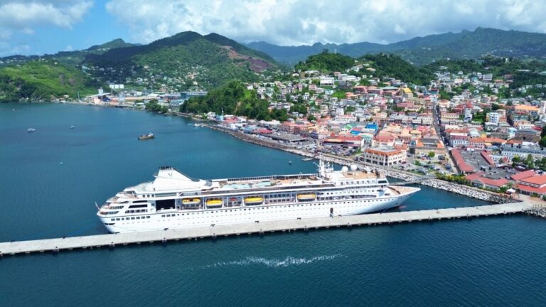 A large cruise ship docked at a port with Villa Vie Residences visible in the background, nestled in a coastal town of colorful buildings and lush green hills. The blue sea and cloudy sky complete this idyllic view.
