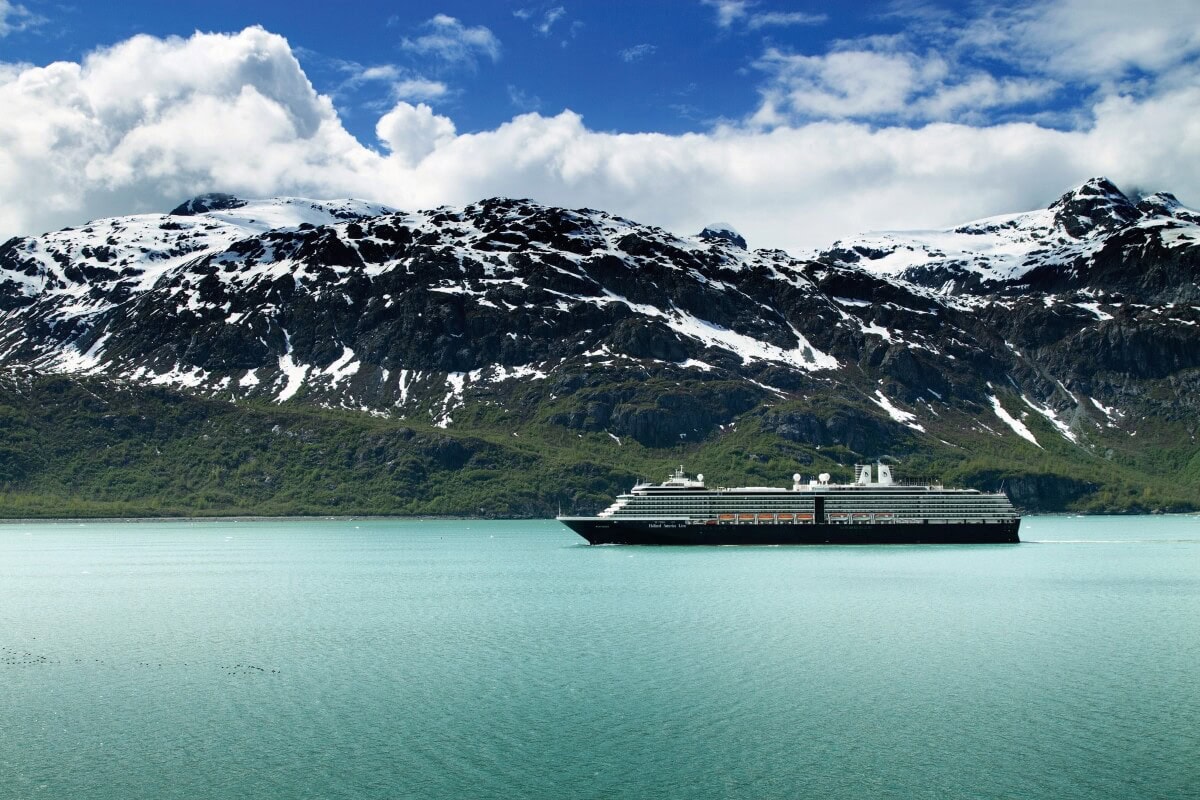 A large Holland America cruise ship sails on a calm turquoise sea with snow-capped mountains in the background under a partly cloudy sky, promising the thrill of free cruises to lucky travelers.