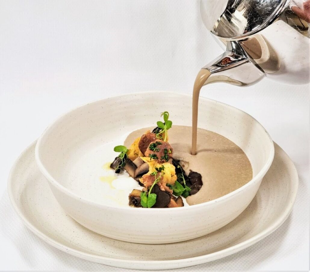 A bowl of creamy mushroom soup, reminiscent of the flavors at Rudi’s Sel de Mer, is poured from a silver pitcher into a white bowl. The soup, featuring bread pieces, mushrooms, and green herbs, rests elegantly on a matching plate against a pristine white background.