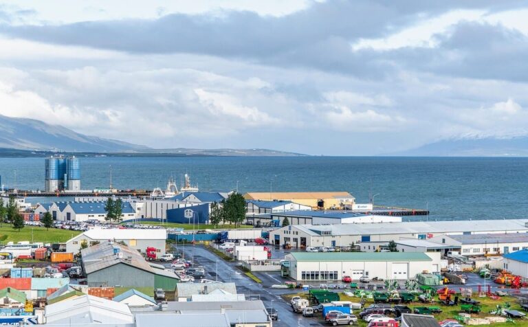 An aerial view of a coastal Icelandic town featuring industrial buildings, warehouses, and scattered residential houses. The sea is to the right, with mountains visible in the distance under a partly cloudy sky. Various vehicles are parked throughout.