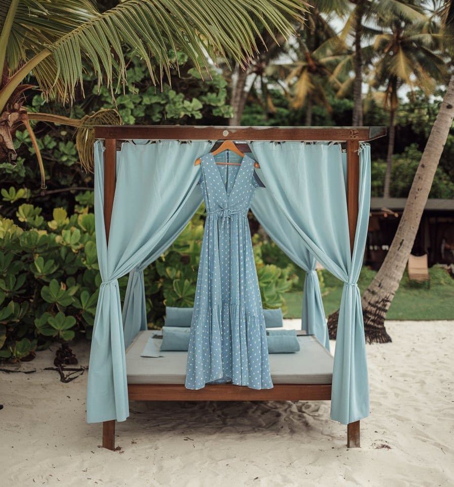 A light blue, sleeveless dress with white polka dots hangs on a wooden canopy bed adorned with matching blue curtains. Perfect for women's cruise fashion, this piece complements a sandy beach setting surrounded by lush green plants and palm trees.
