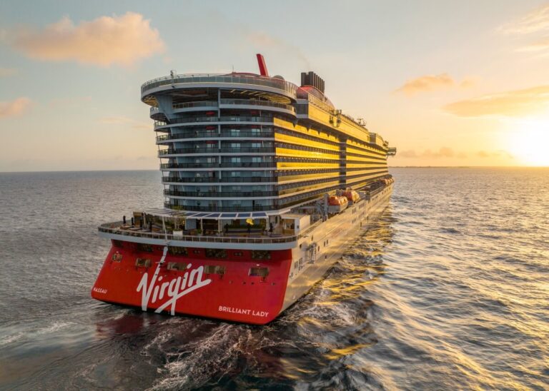 The majestic Virgin Brilliant Lady from Virgin Voyages gracefully sails across the ocean during sunset. This luxurious cruise ship, adorned in red and white, glides effortlessly as the sky bursts with warm tones, offering a stunning backdrop for those enjoying their annual pass upgrades.