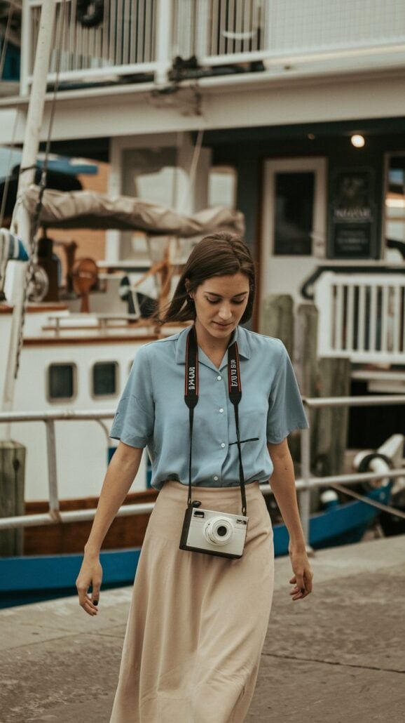 A woman strolls along a dock with boats in the background, embracing the vibe of her Hawaiian cruise. She's dressed in a blue blouse and beige skirt, with a camera—the perfect addition to her packing list—hanging around her neck.
