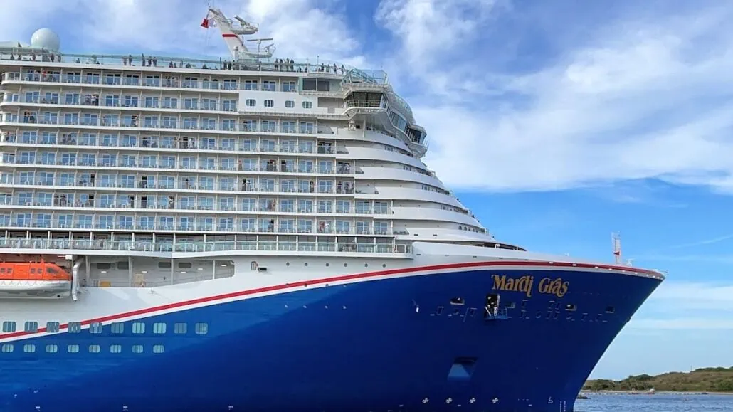 Side view of the large cruise ship "Mardi Gras" with its bright blue hull and multiple decks with balconies. On this busiest day at Port Canaveral, the sky is partly cloudy, and greenery is visible in the distance as 25 vessels fill the open waters where it is docked.