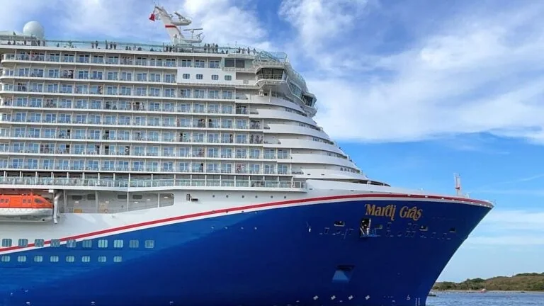 Side view of the large cruise ship "Mardi Gras" with its bright blue hull and multiple decks with balconies. On this busiest day at Port Canaveral, the sky is partly cloudy, and greenery is visible in the distance as 25 vessels fill the open waters where it is docked.