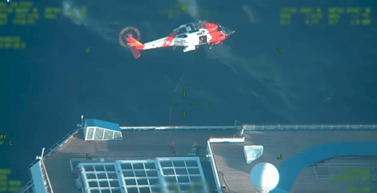 Aerial view of a red and white helicopter hovering over Carnival Valor at sea. The Coast Guard is airlifting a passenger from the vessel's deck using a rescue line. The ship features a large glass structure and a circular white object.