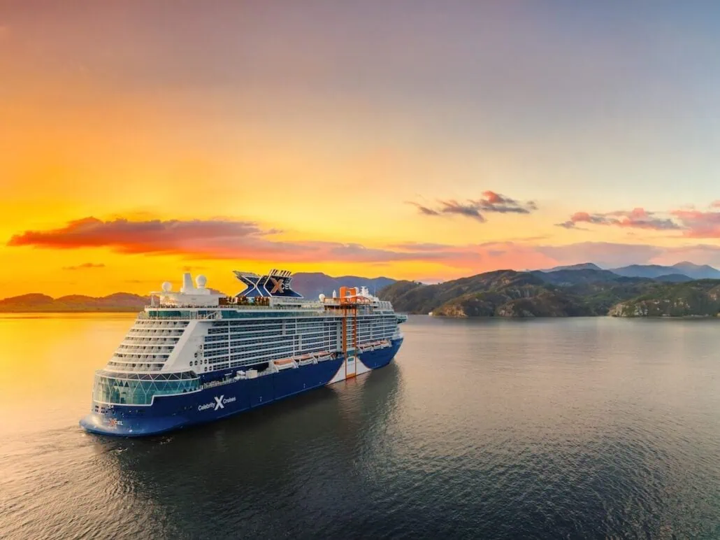 A Celebrity Cruises ship gracefully sails on calm water at sunset. The sky is a beautiful tapestry of orange and pink, with a few clouds. Hills or mountains silhouette the horizon, while the ship's stylish design elevates its logo above. The water mirrors the warm colors of this serene scene.