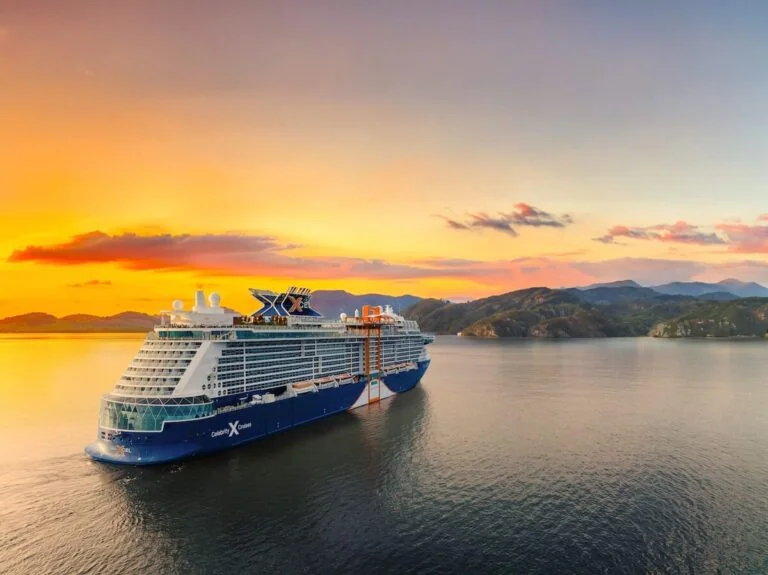 A Celebrity Cruises ship gracefully sails on calm water at sunset. The sky is a beautiful tapestry of orange and pink, with a few clouds. Hills or mountains silhouette the horizon, while the ship's stylish design elevates its logo above. The water mirrors the warm colors of this serene scene.