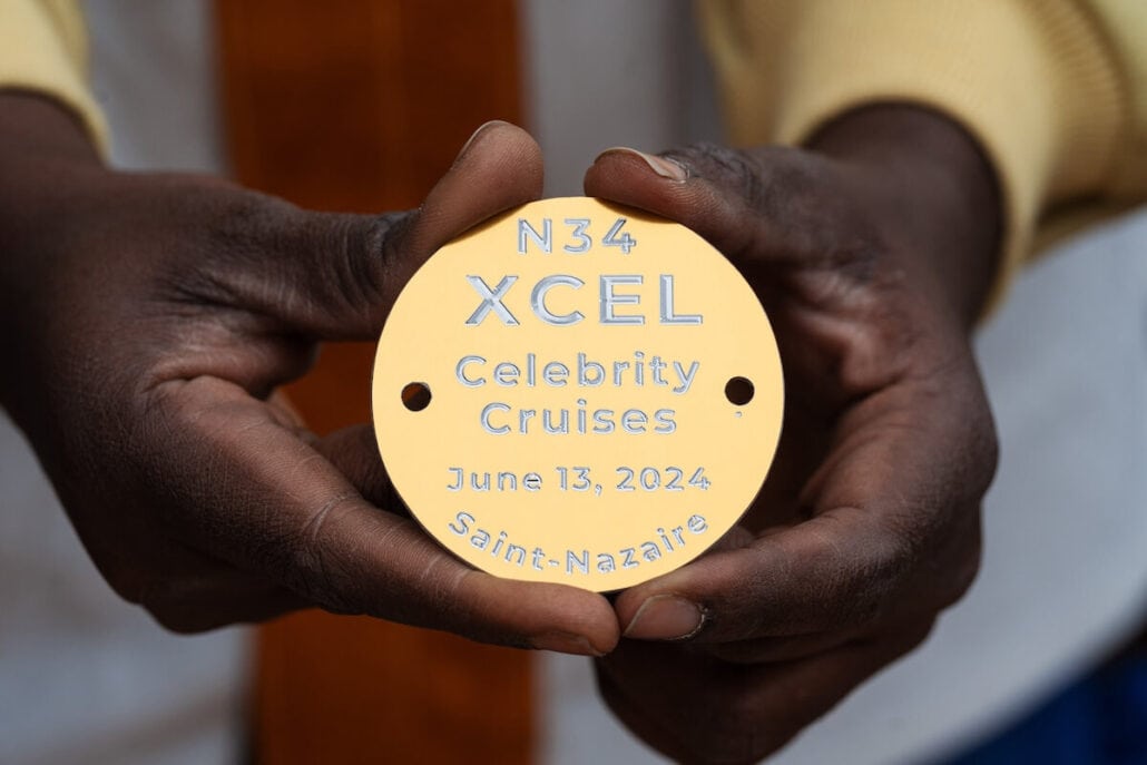 Hands holding a round, gold-colored plaque engraved with "N34 XCEL Celebrity Cruises June 13, 2024 Saint-Nazaire," marking the launch of the Design Next Ship, poised to delight future passengers with unparalleled luxury and innovation.