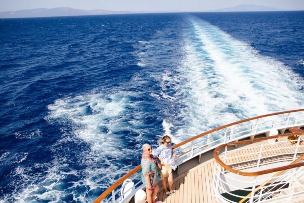 A man and a woman stand together on the deck of a Celestyal Cruises ship, looking out at the Arabian Gulf. The vessel leaves a trail in the blue water. The sky is clear, and distant land is visible on the horizon as their itinerary unfolds.
