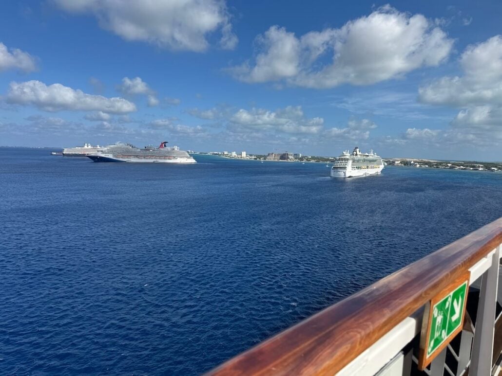 From the ship's deck, gaze out at the ocean beneath a partly cloudy sky. In the distance, two cruise ships linger, hinting at a decline in their numbers near the Cayman Islands. The coastline and city skyline on the horizon complete this picturesque view framed by a safety sign and railing.
