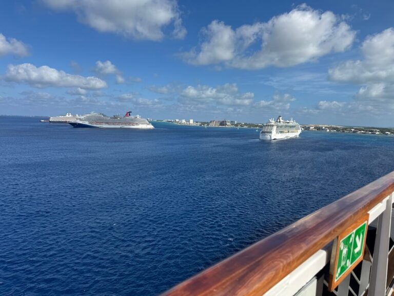 From the ship's deck, gaze out at the ocean beneath a partly cloudy sky. In the distance, two cruise ships linger, hinting at a decline in their numbers near the Cayman Islands. The coastline and city skyline on the horizon complete this picturesque view framed by a safety sign and railing.