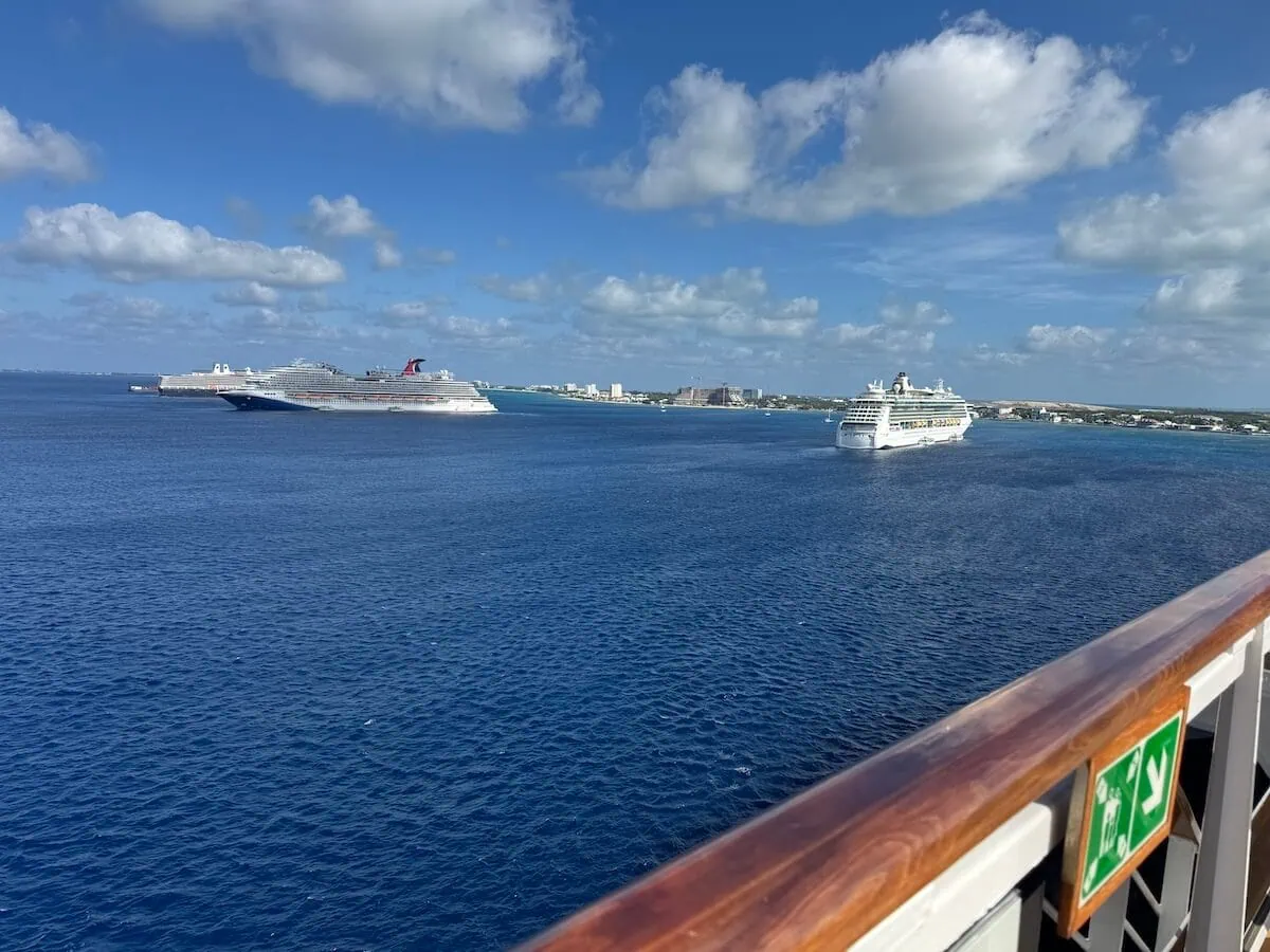 From the ship's deck, gaze out at the ocean beneath a partly cloudy sky. In the distance, two cruise ships linger, hinting at a decline in their numbers near the Cayman Islands. The coastline and city skyline on the horizon complete this picturesque view framed by a safety sign and railing.