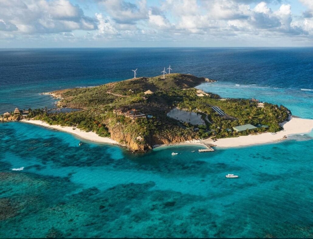 Aerial view of a small tropical island surrounded by turquoise water. The island features lush greenery, sandy beaches, several buildings, and three wind turbines that received significant upgrades. A few boats are visible in the water near the shore.